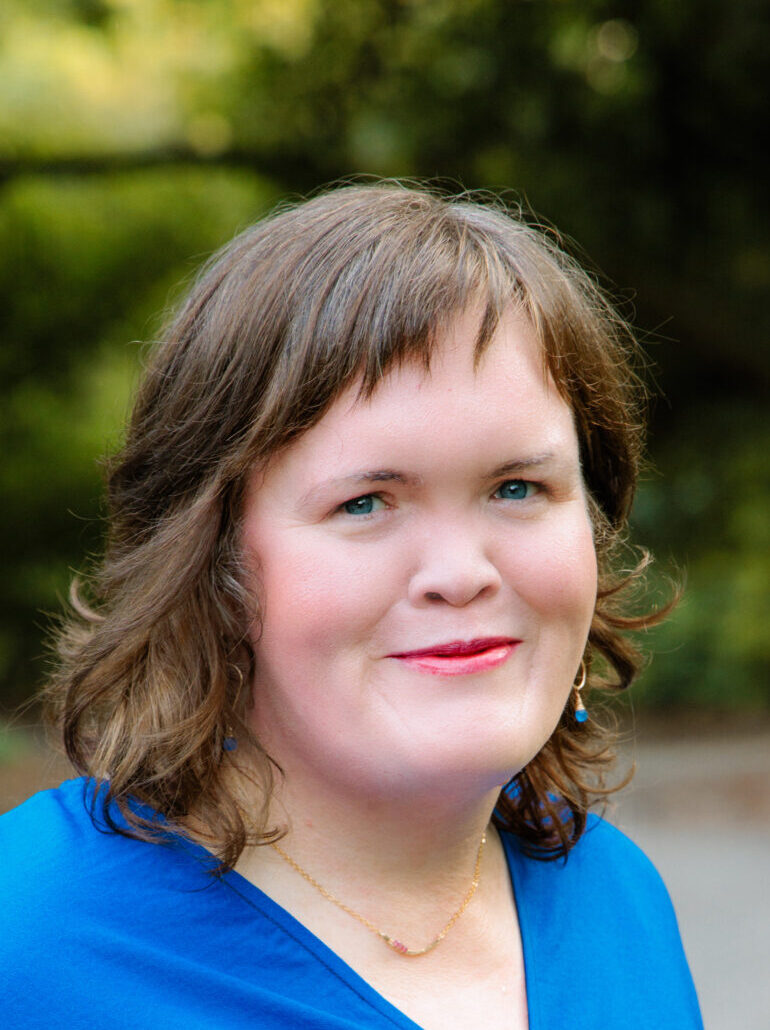 Carrie in a bright blue dress in the woods smiling with closed lips